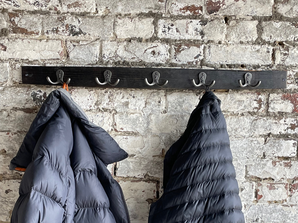 Black coat rack with five hooks mounted on a weathered brick wall, holding two dark puffer jackets.