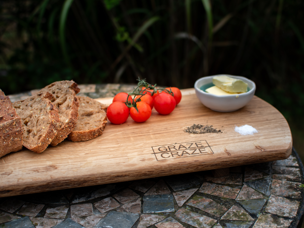 A wooden grazing board with bread, tomatoes, salt, pepper and butter in a ceramic pot on it.
