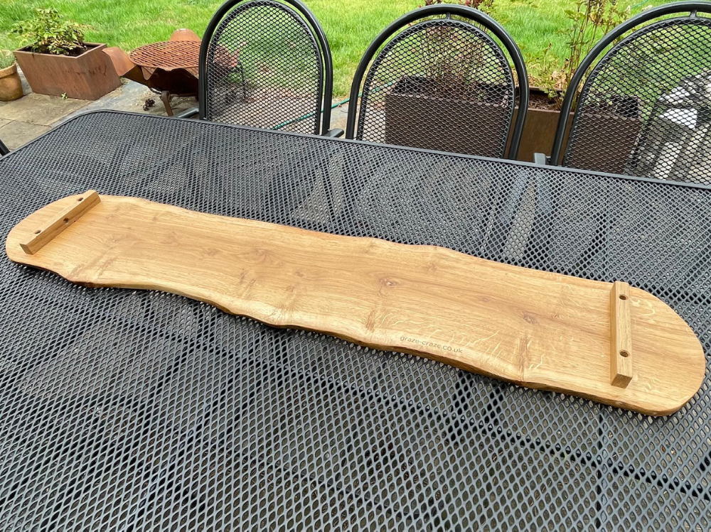 A reverse view of a long, wooden grazing board with natural edges sits on a black metal table outdoors. Two small wooden legs are on each end.