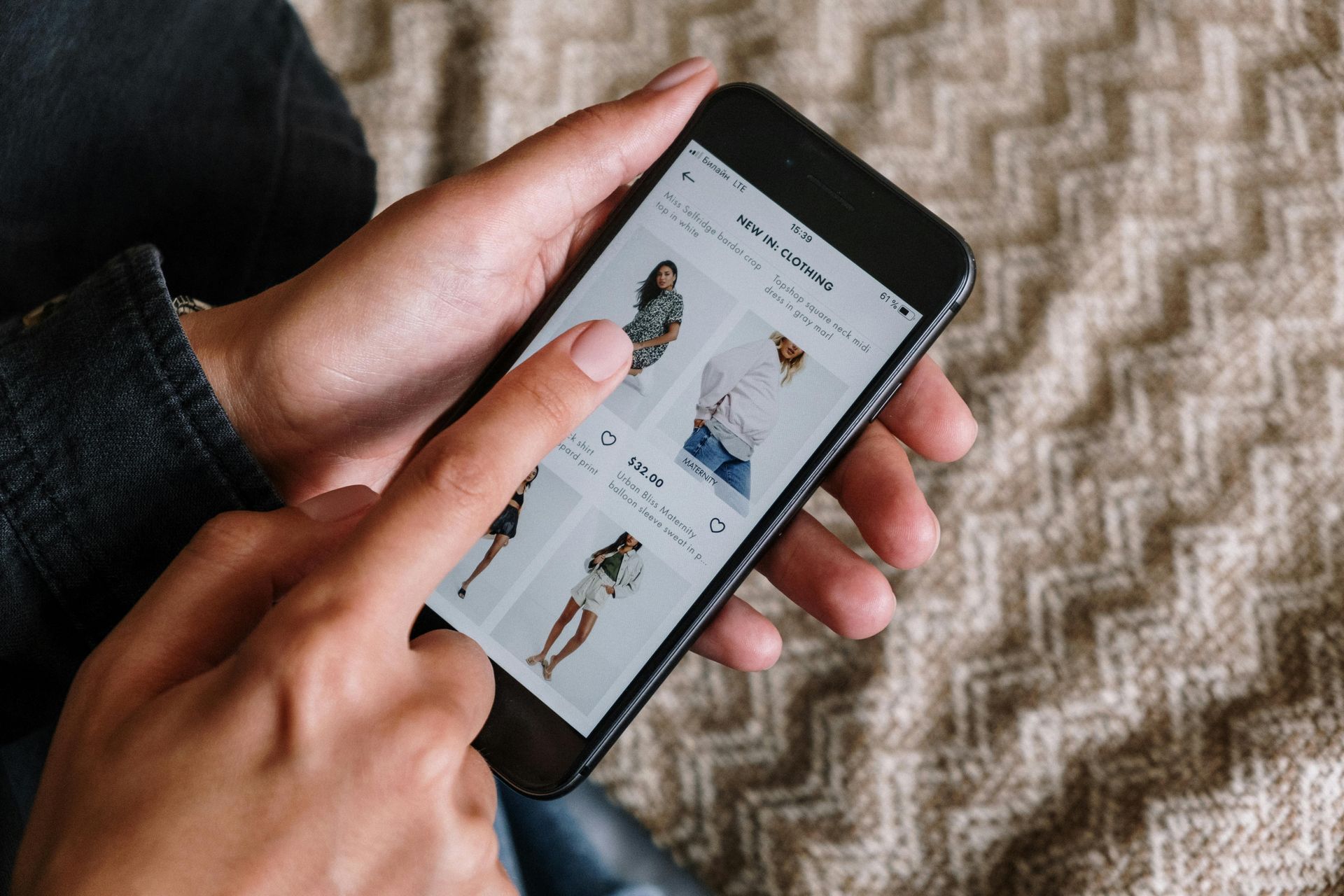 Person using a smartphone to shop online for clothing; fingers tap on screen showing product images.