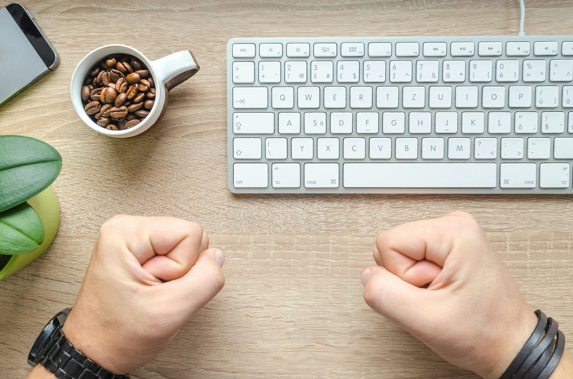 Hands clenched in fists on a wooden desk with a keyboard, coffee cup, phone, and plant.