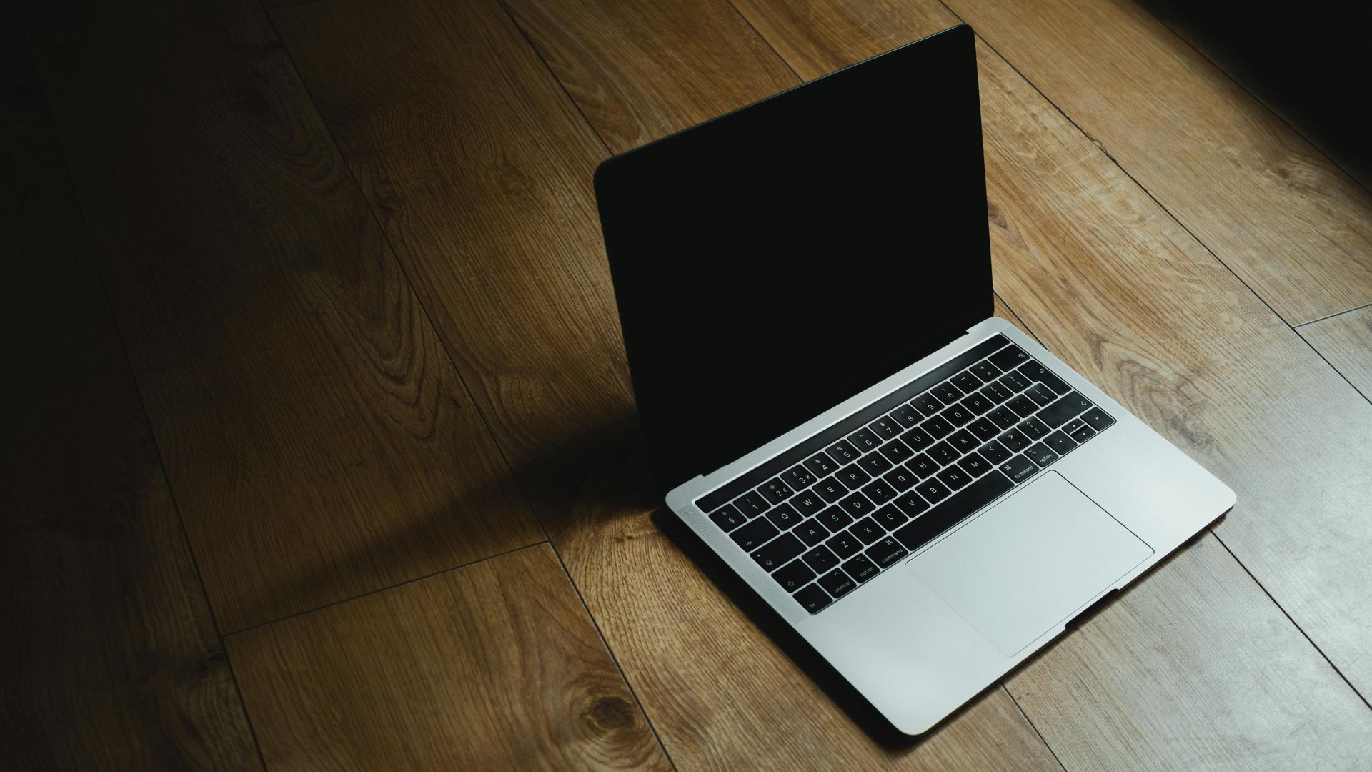 Laptop on a wooden floor, screen open, casting a shadow in a dimly lit room.
