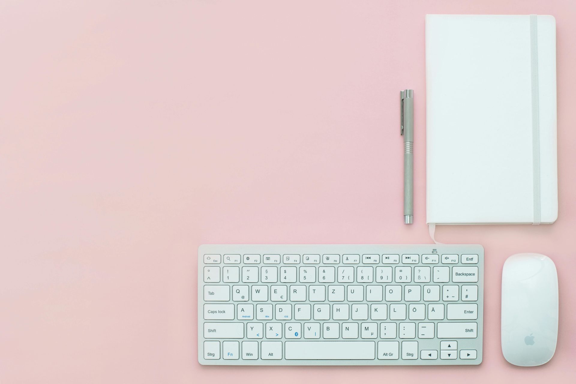 White keyboard, mouse, notebook, and pen on a light pink surface.