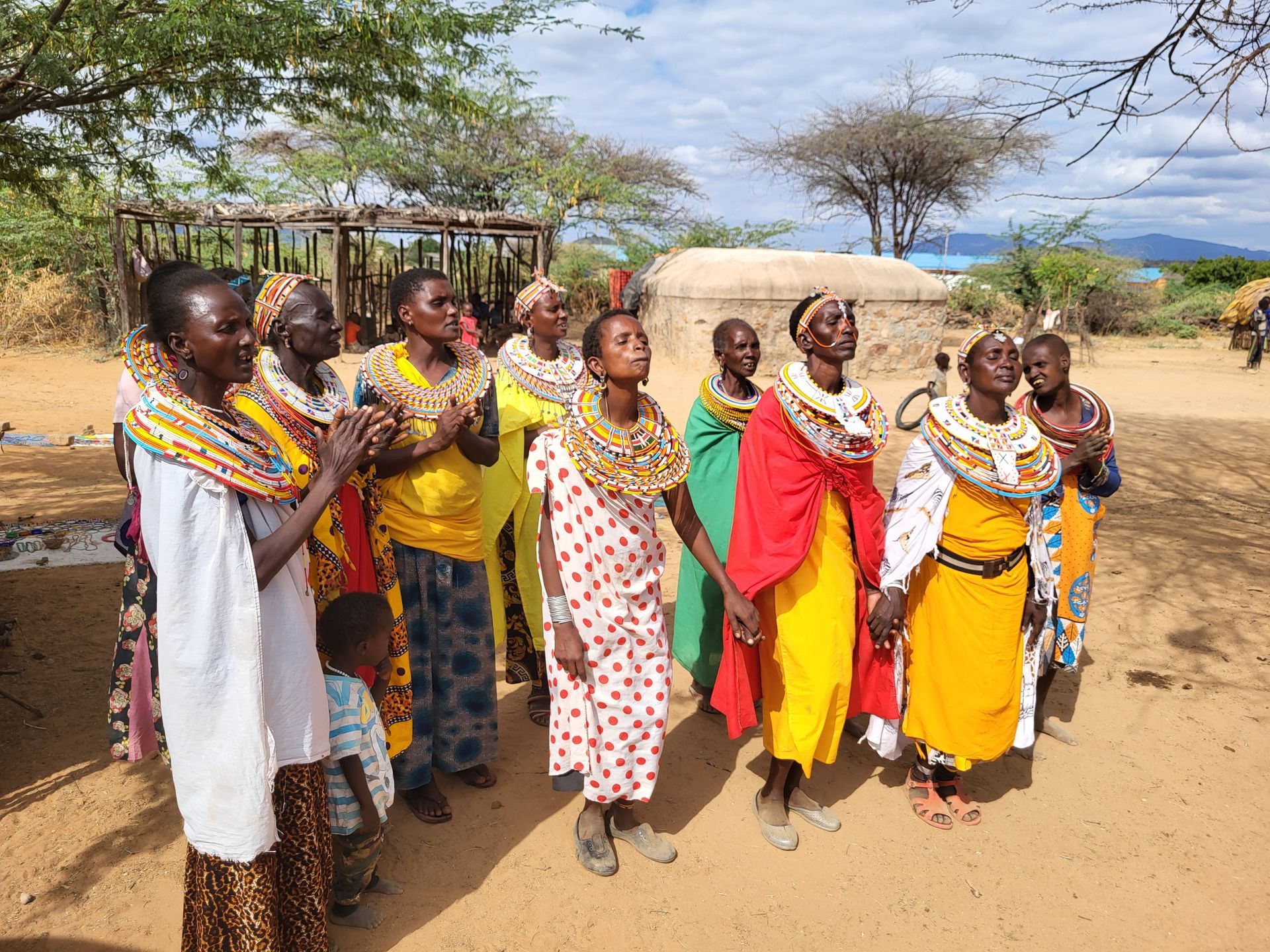 Group of people in colorful clothing, some with beaded necklaces, outdoors, looking upwards.