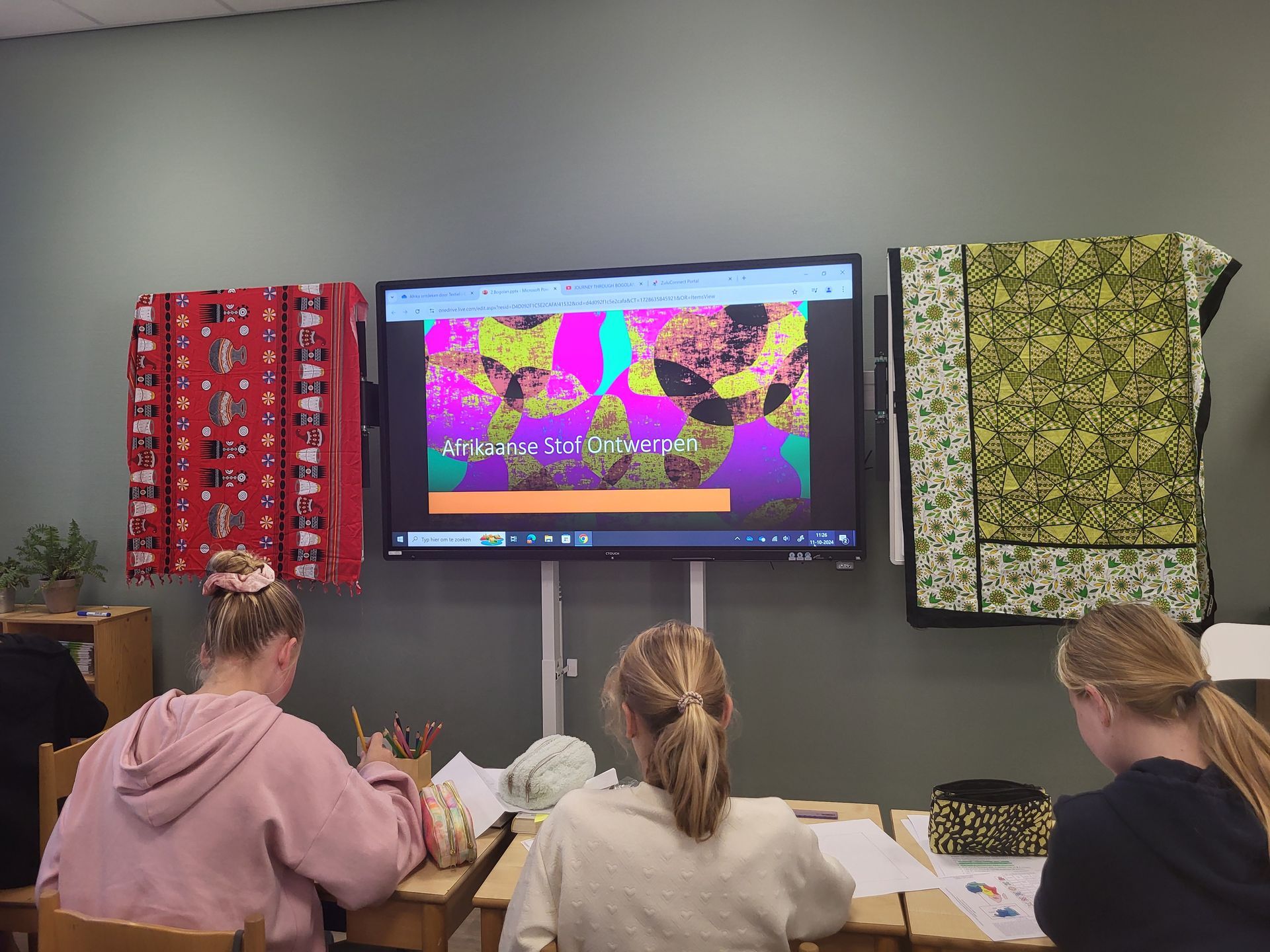 Three students at desks watching a screen showing colorful graphics. Quilts hang on the wall.