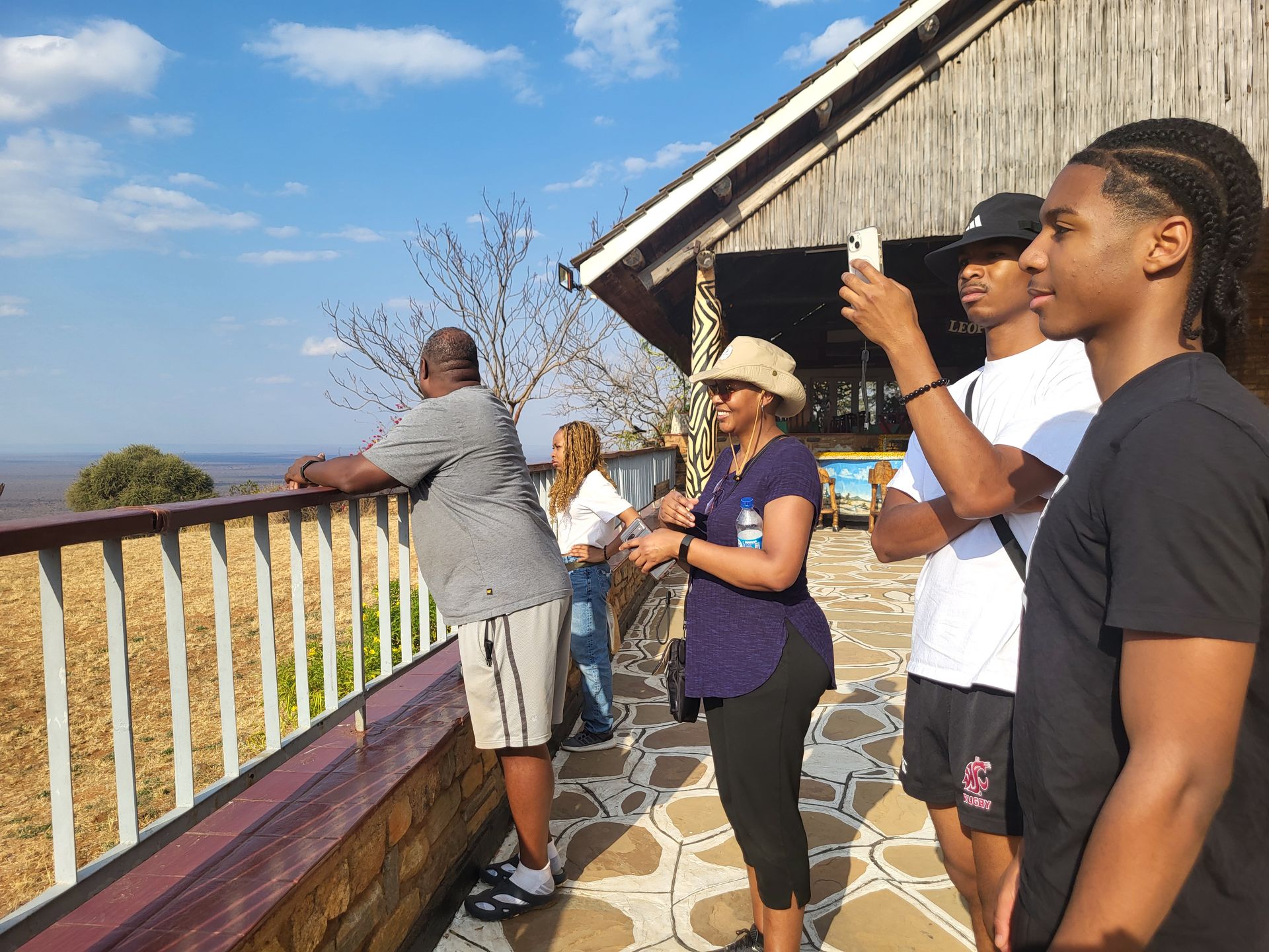 People at a lookout point, some taking photos. A wooden building and blue sky in the background.