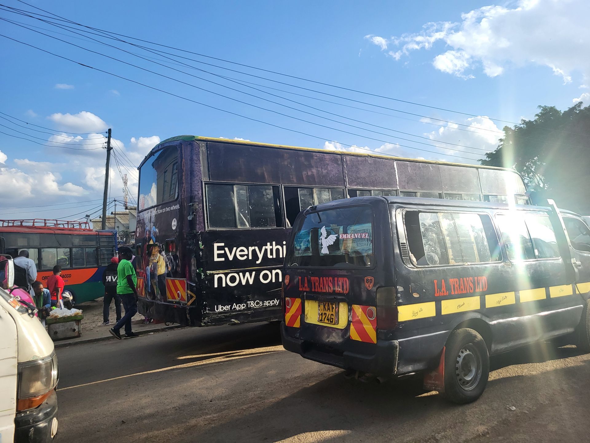 Double-decker bus and minibus on a road in Nairobi, Kenya. Vehicles are black with colorful markings. People stand nearby.