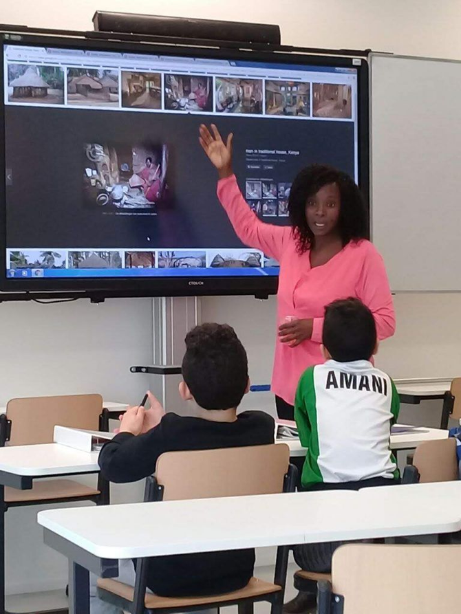 Teacher pointing at a screen showing images to two students in a classroom. The teacher is wearing pink.