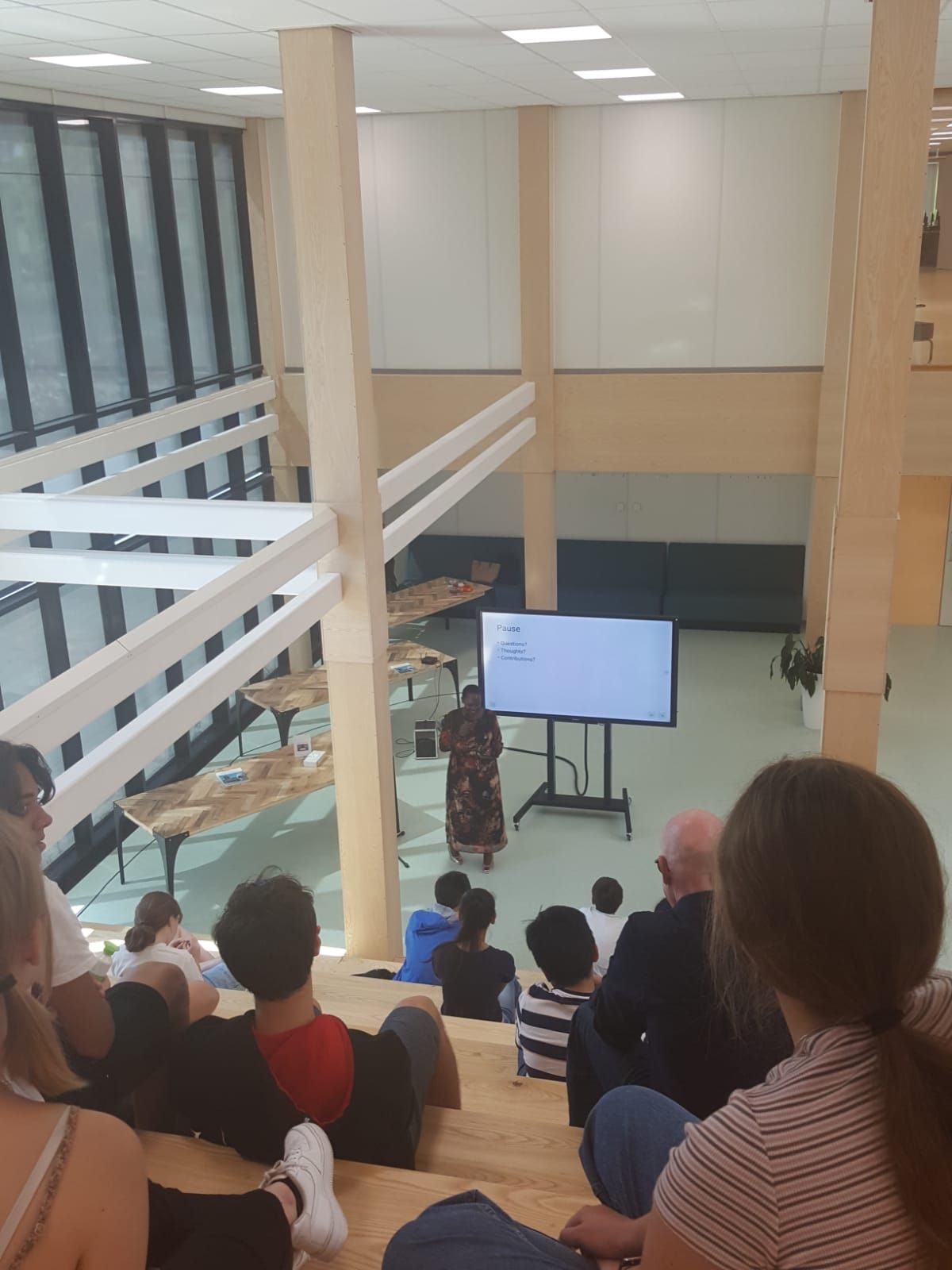 Person presenting to a seated audience in a multi-level open space. A screen displays a presentation.