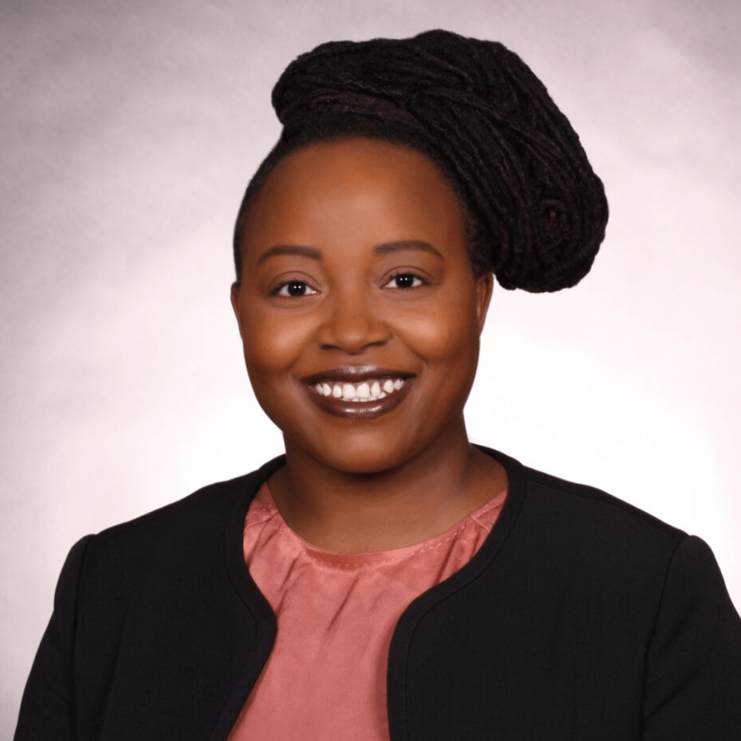 Woman smiling, wearing a black blazer over a pink blouse; hair styled in dark braids.