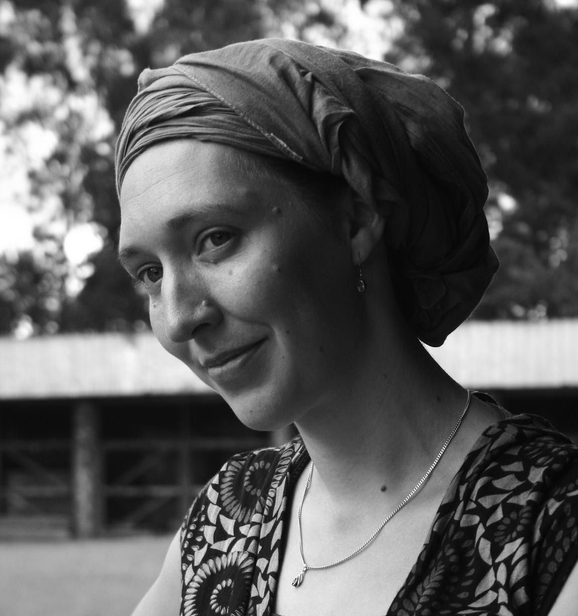 Woman wearing a headscarf, smiling, looking to the side. Wearing a patterned top and necklace.