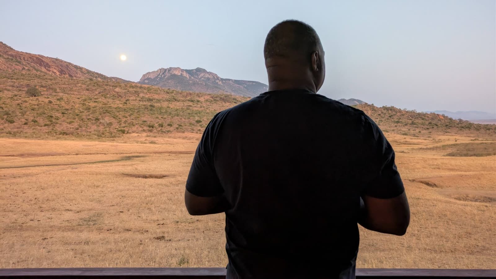 Man overlooking a vast savanna landscape; mountains in the distance, moon in the sky.
