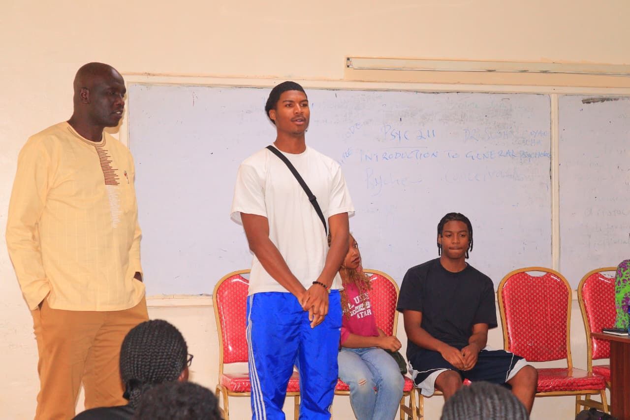 People in a room, with a man speaking, others seated.  Whiteboard in background.