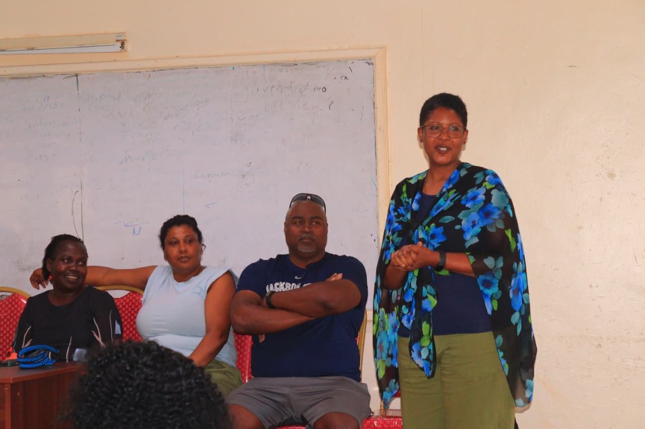 A woman speaks to an audience; three seated people listen. Room with whiteboard.