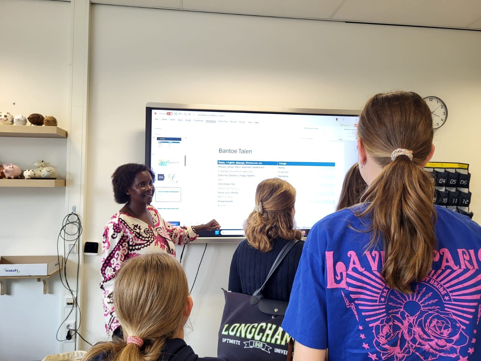Woman points at screen, speaking to group of students in a classroom.