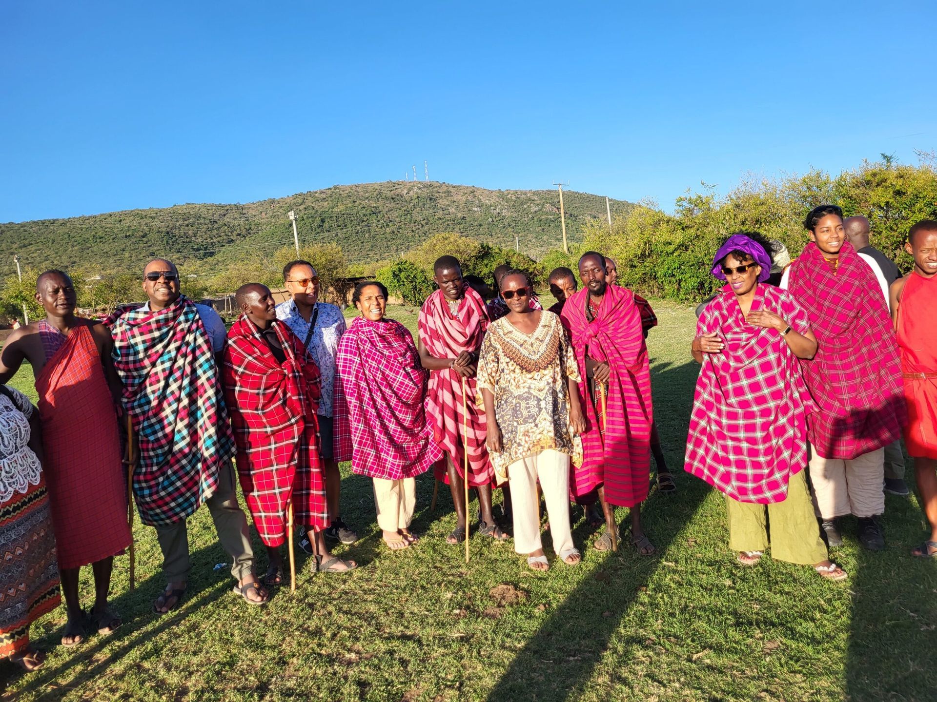 Group of people in traditional attire pose outdoors, with a hill and blue sky background.