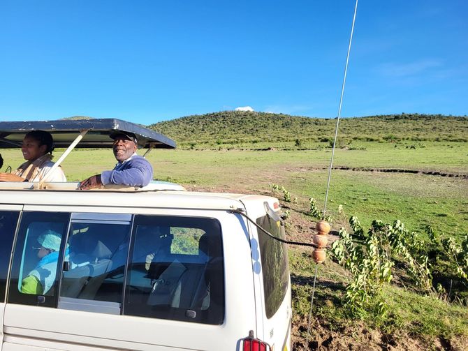People in a safari vehicle driving on a dirt road, grassy field and a hilltop in the background.
