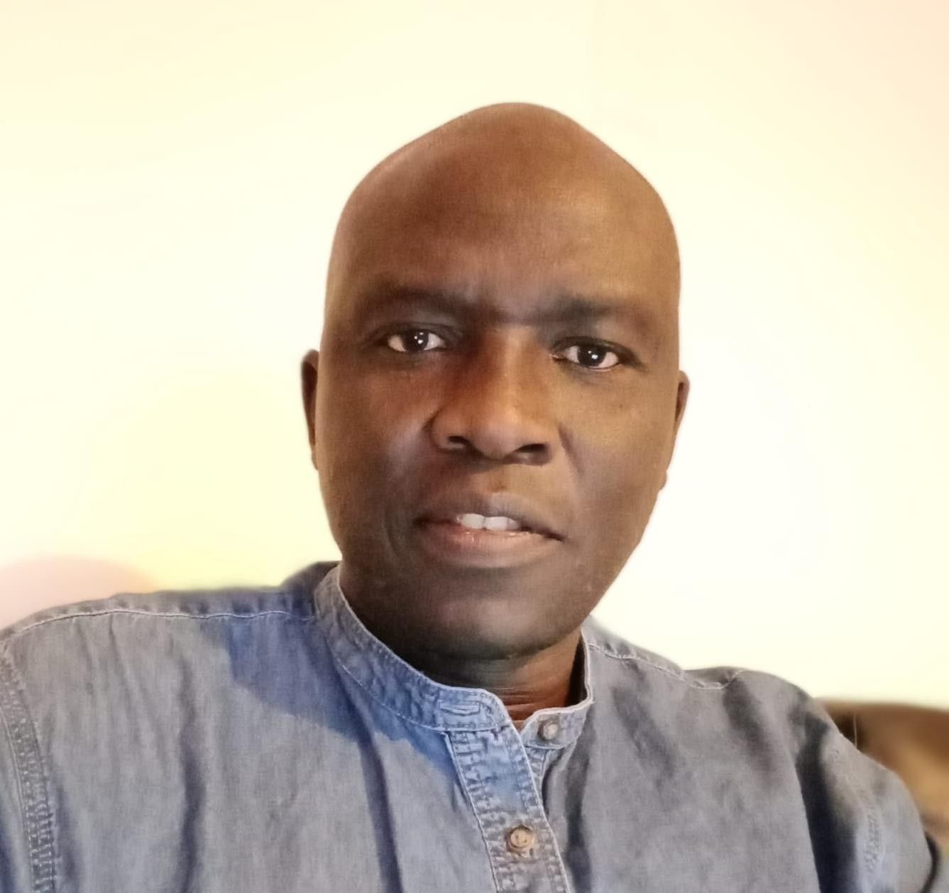 Man with a shaved head wearing a blue denim shirt against a neutral background.