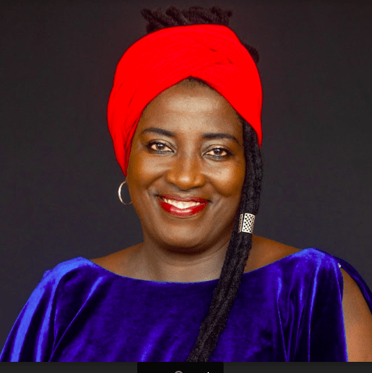Woman wearing a red head wrap and blue velvet dress smiles at the camera against a dark background.