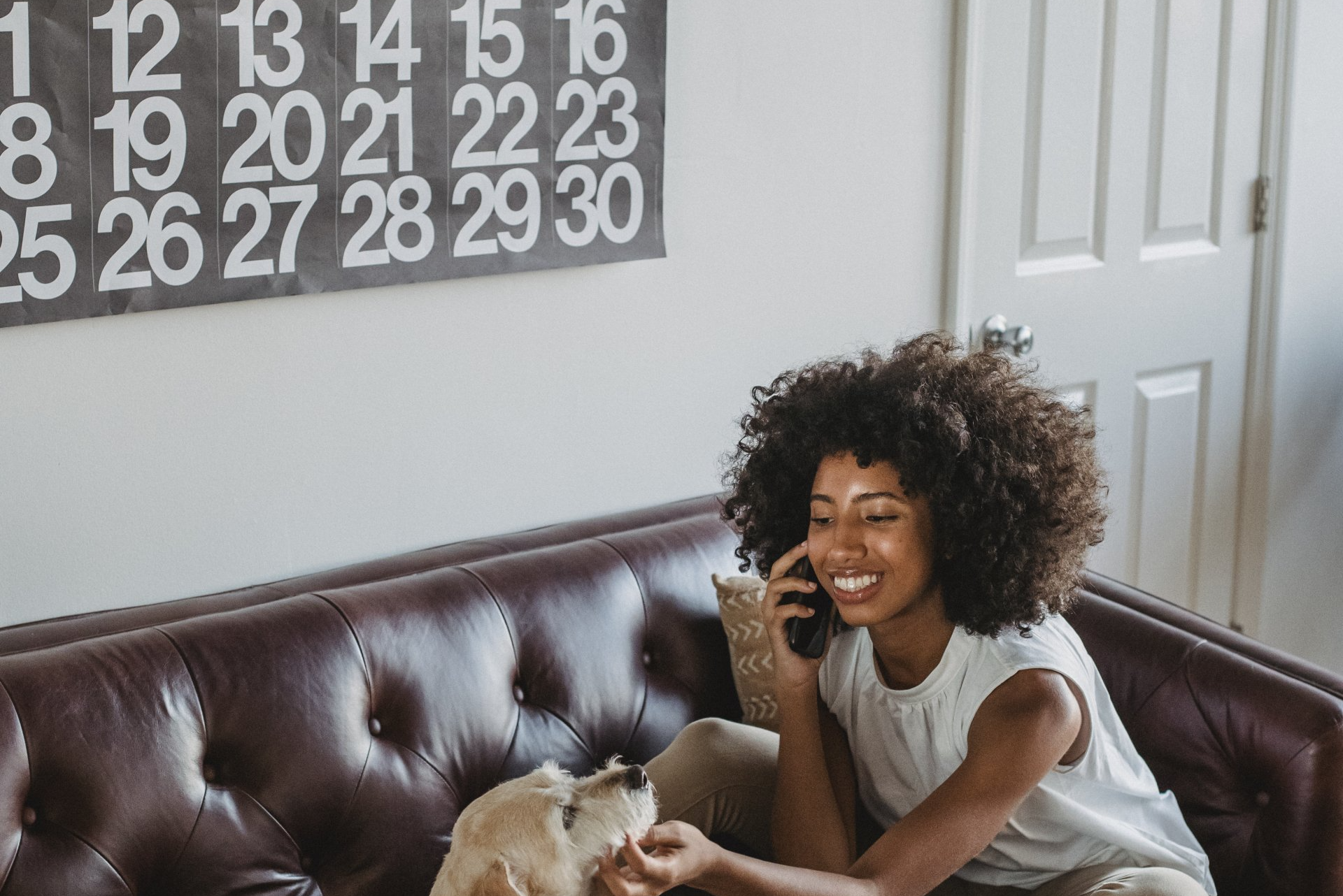 A woman is sitting on a couch talking on a cell phone while petting her dog.