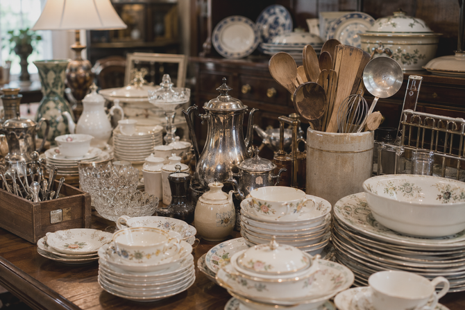 Table filled with stacked white dishes, glassware, and serving utensils in a cozy antique shop display