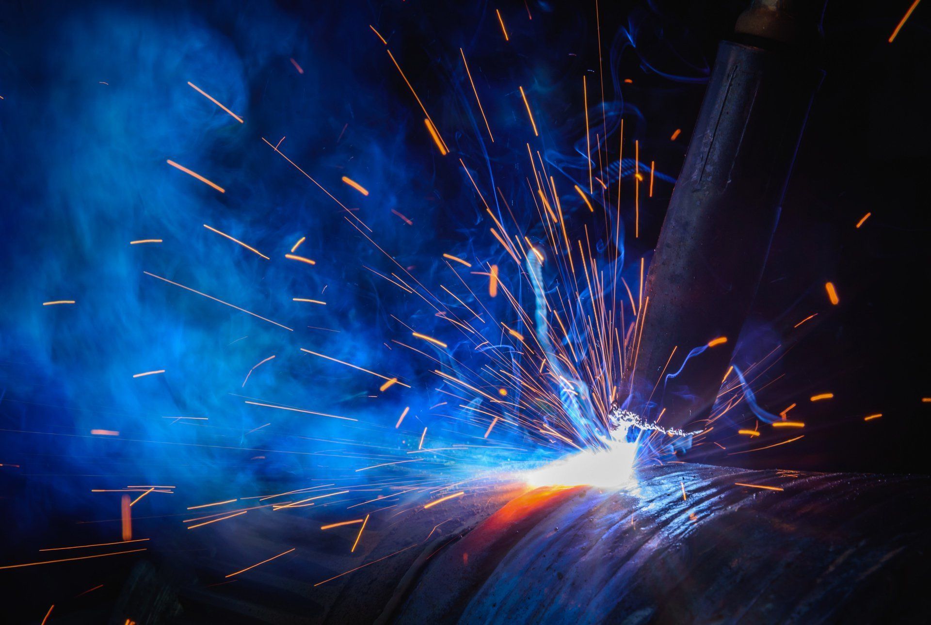 A welding torch creates a bright white glow and orange sparks against a dark background with blue smoke.