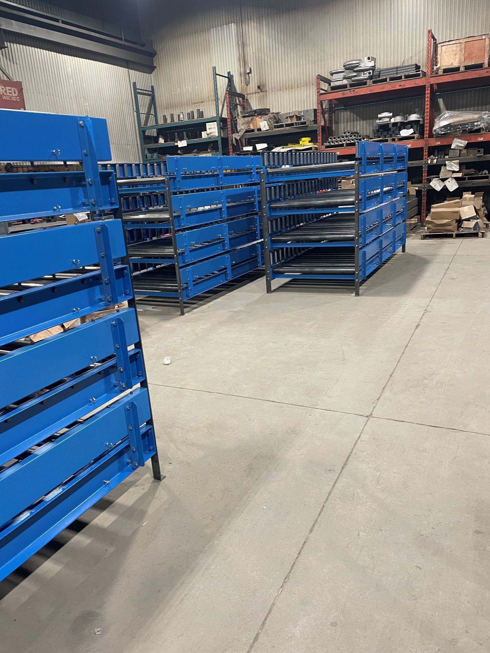 Rows of blue industrial storage racks in a warehouse with shelving units filled with various parts in the background.