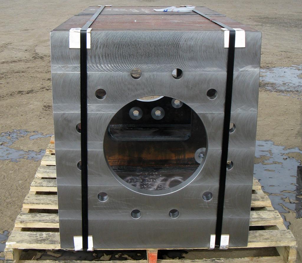 A heavy, rectangular steel block with a large circular hole and multiple bolt holes, strapped to a wooden pallet.