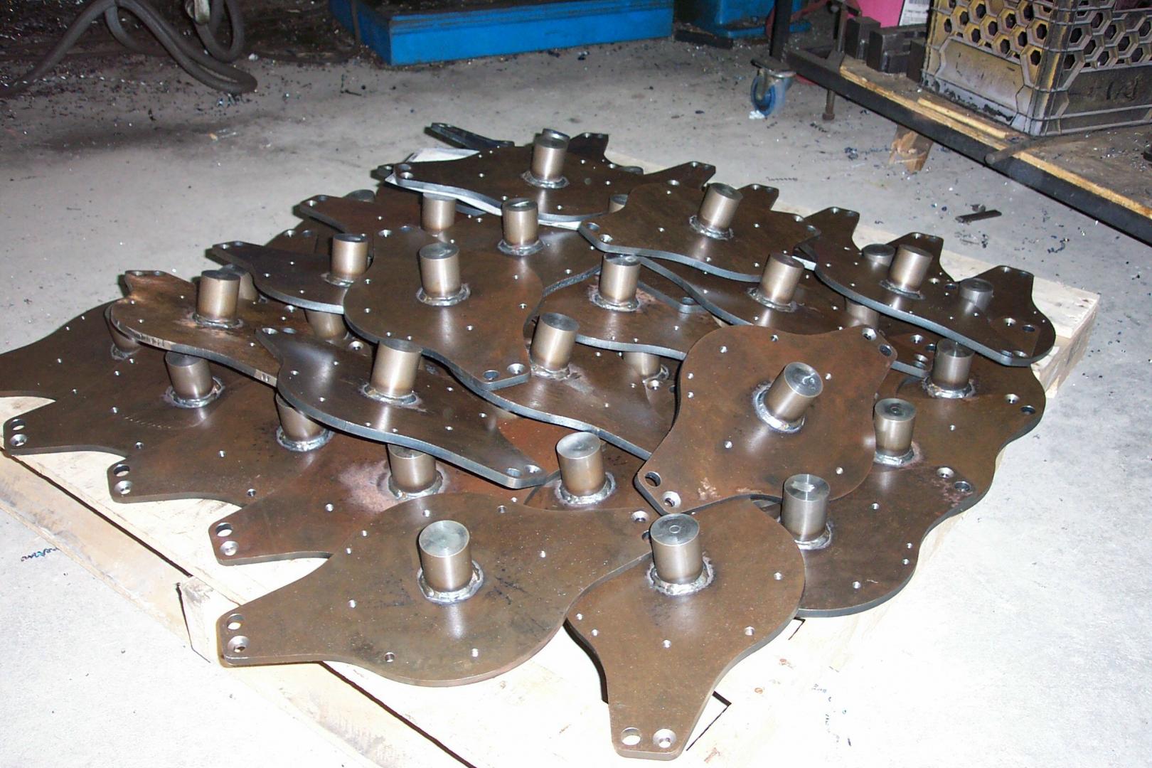 A pile of industrial metal plates with cylindrical hubs welded to their centers, stacked on a wooden pallet in a workshop.
