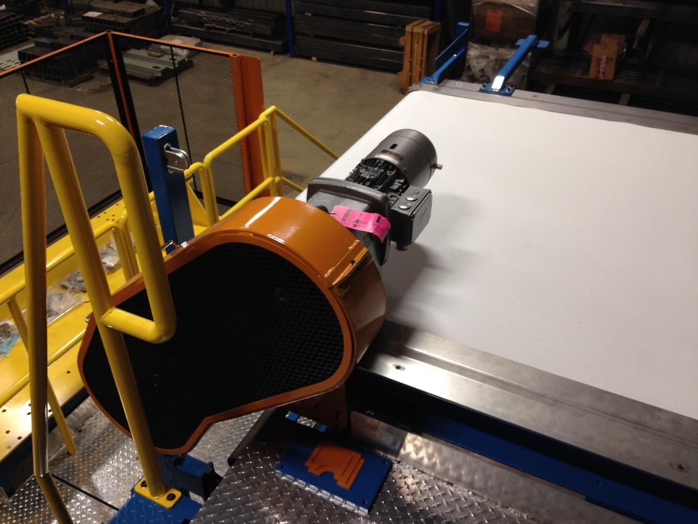 A yellow safety railing stands next to an industrial machine with a bright orange housing and a gray motor.