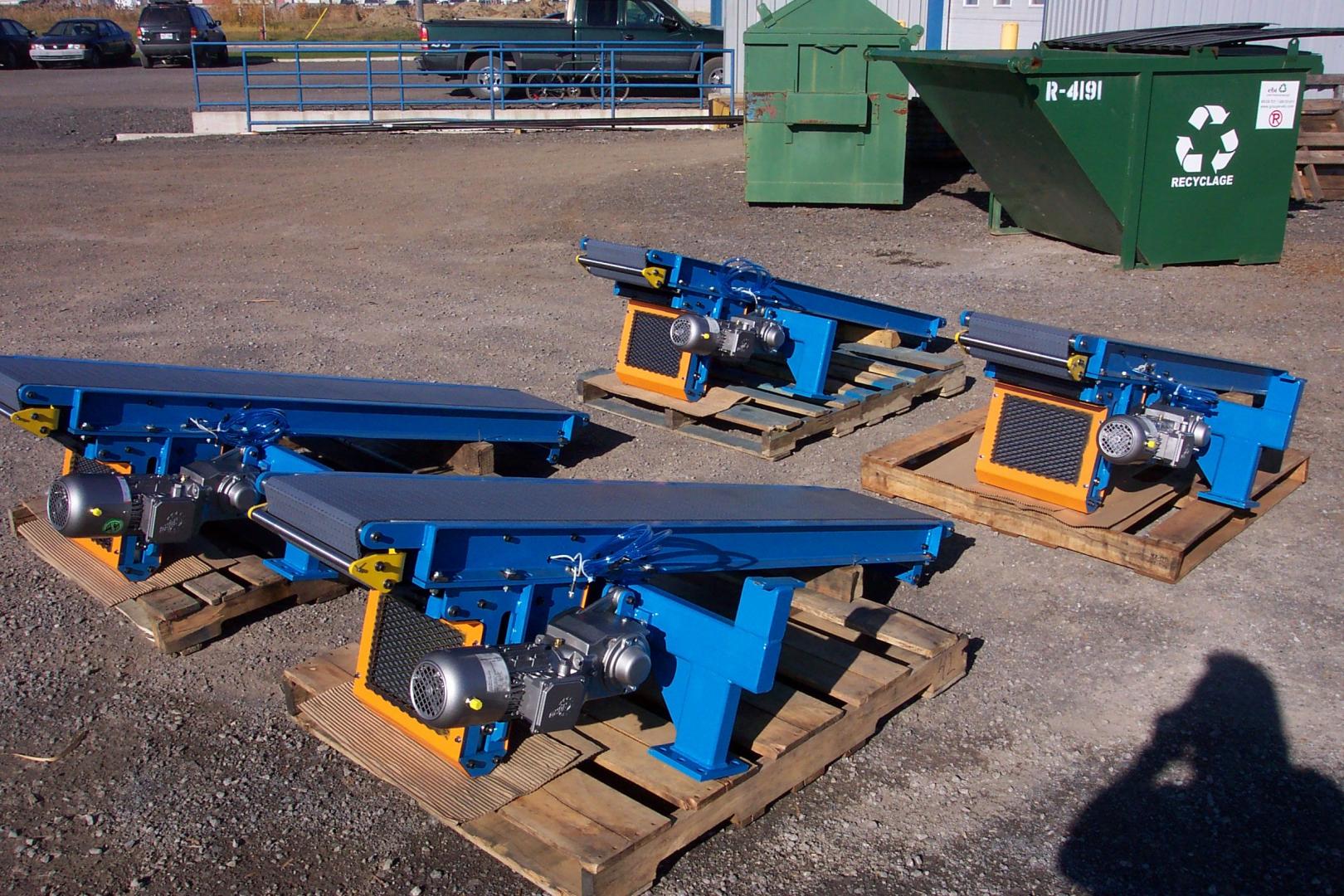 Three blue industrial vibratory conveyor feeders on wooden pallets sit in a paved outdoor lot near green dumpsters.