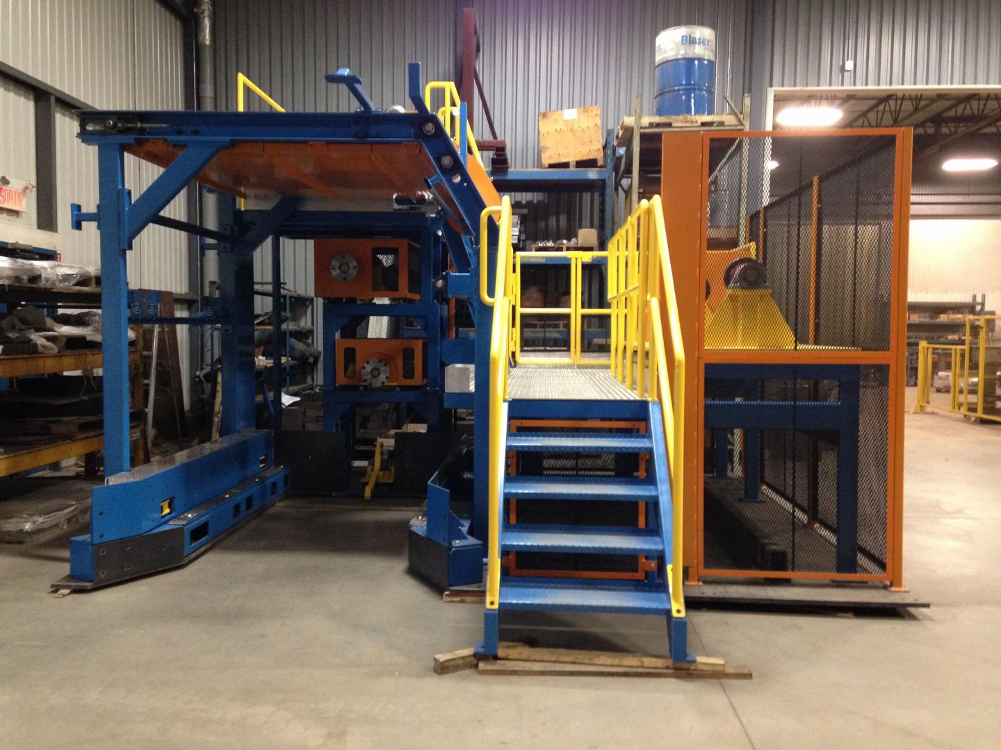 Large industrial machine in a warehouse with a blue frame, orange platform, yellow metal stairs, and a screened area.