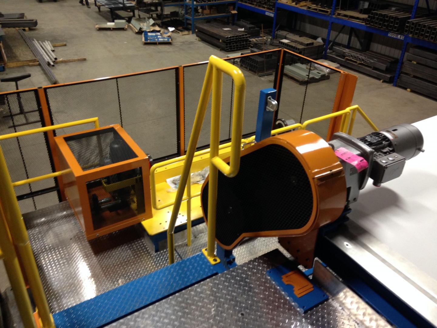 A high-angle view of industrial machinery with bright orange safety guards and yellow railings on a metal platform.