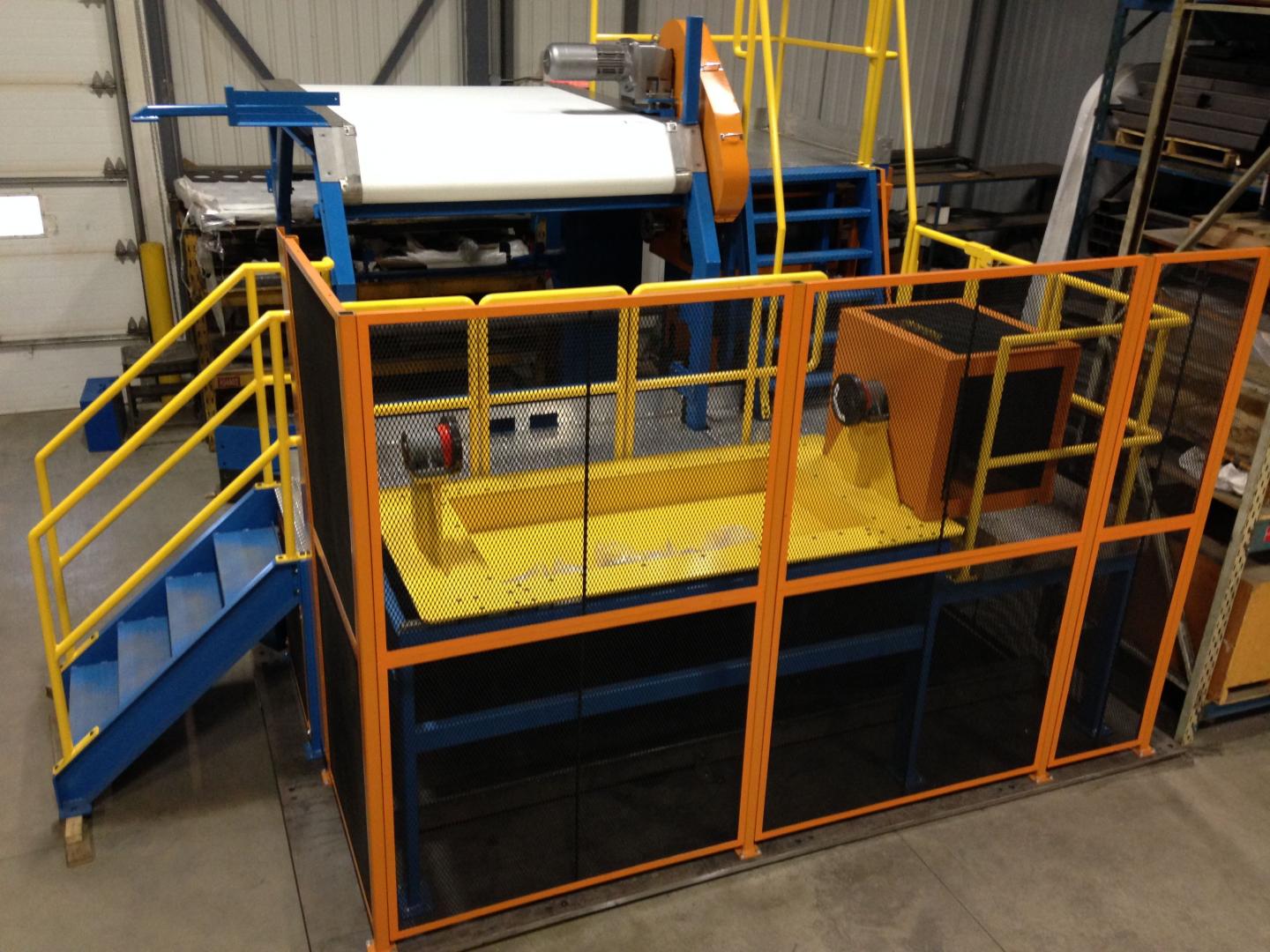 Industrial machine with a yellow grated platform, safety railings, and a blue staircase in a warehouse setting.