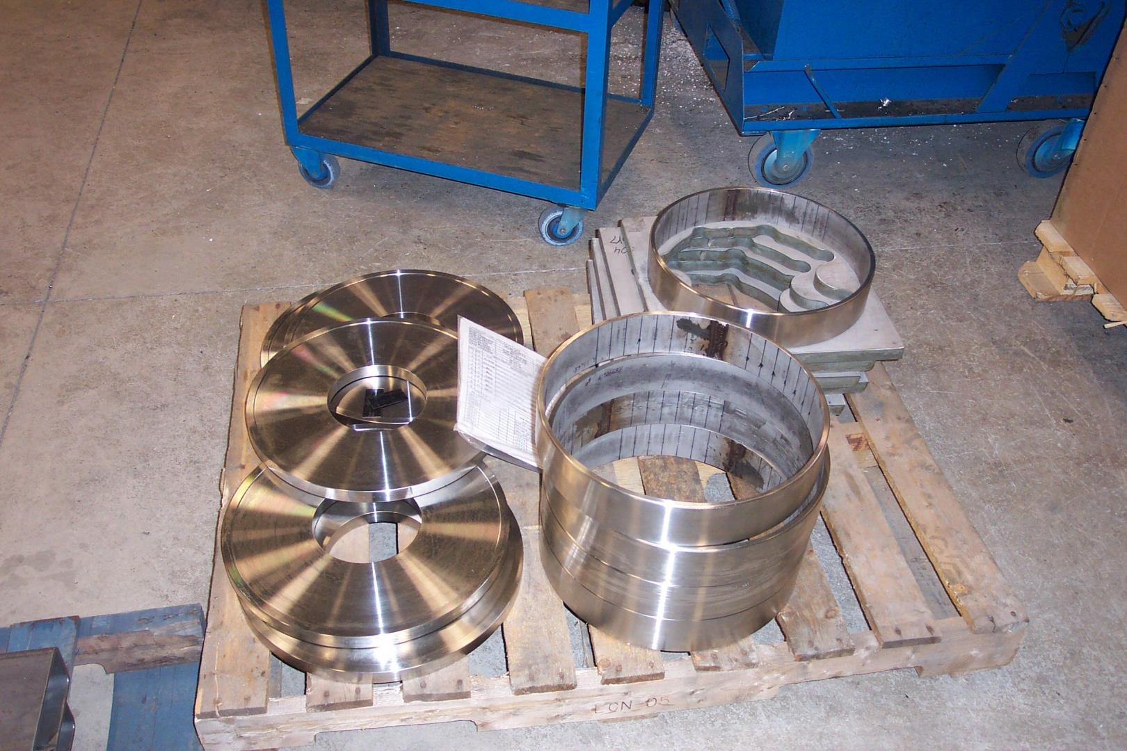 Des anneaux métalliques industriels et des composants circulaires plats reposent sur une palette en bois, elle-même posée sur le sol en béton d'un entrepôt.