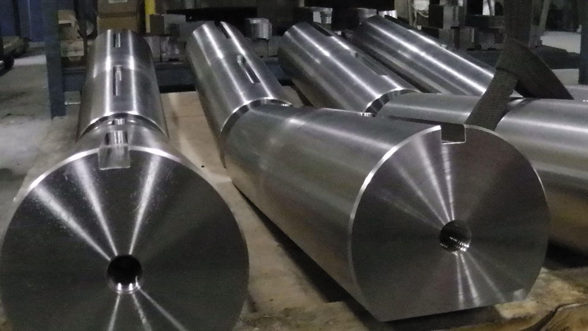 Trois grands arbres industriels cylindriques en métal, munis de rainures de clavette et de trous percés au centre, reposent sur une palette d'atelier.