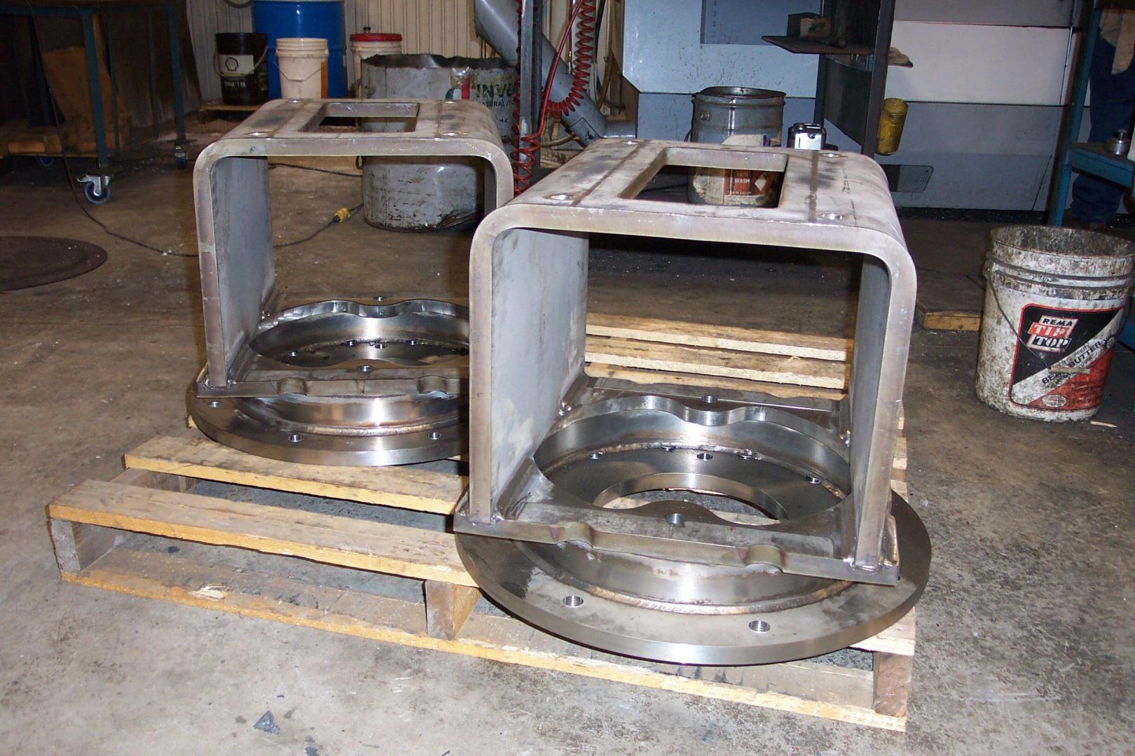 Two C-shaped steel industrial frames mounted on circular base plates, sitting on a wooden pallet in a workshop.