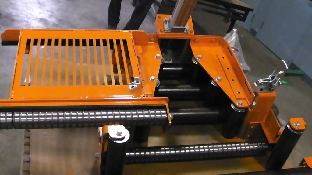 An orange metal industrial conveyor assembly with horizontal rollers and a protective grate, viewed from above.