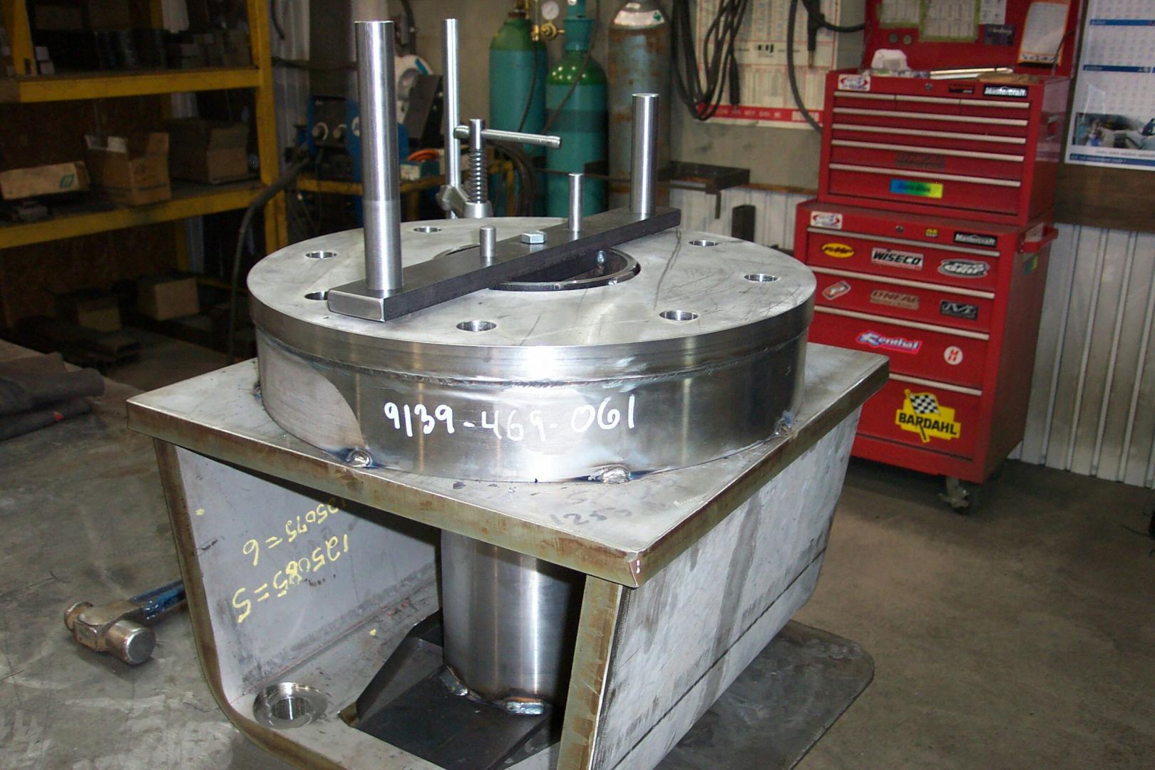 A large, circular steel component with attached guide pins sits on a metal stand in a machine shop.