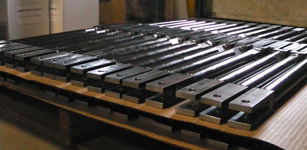 Rows of long, rectangular, dark metal parts with silver brackets stacked neatly on a wooden pallet in a warehouse.