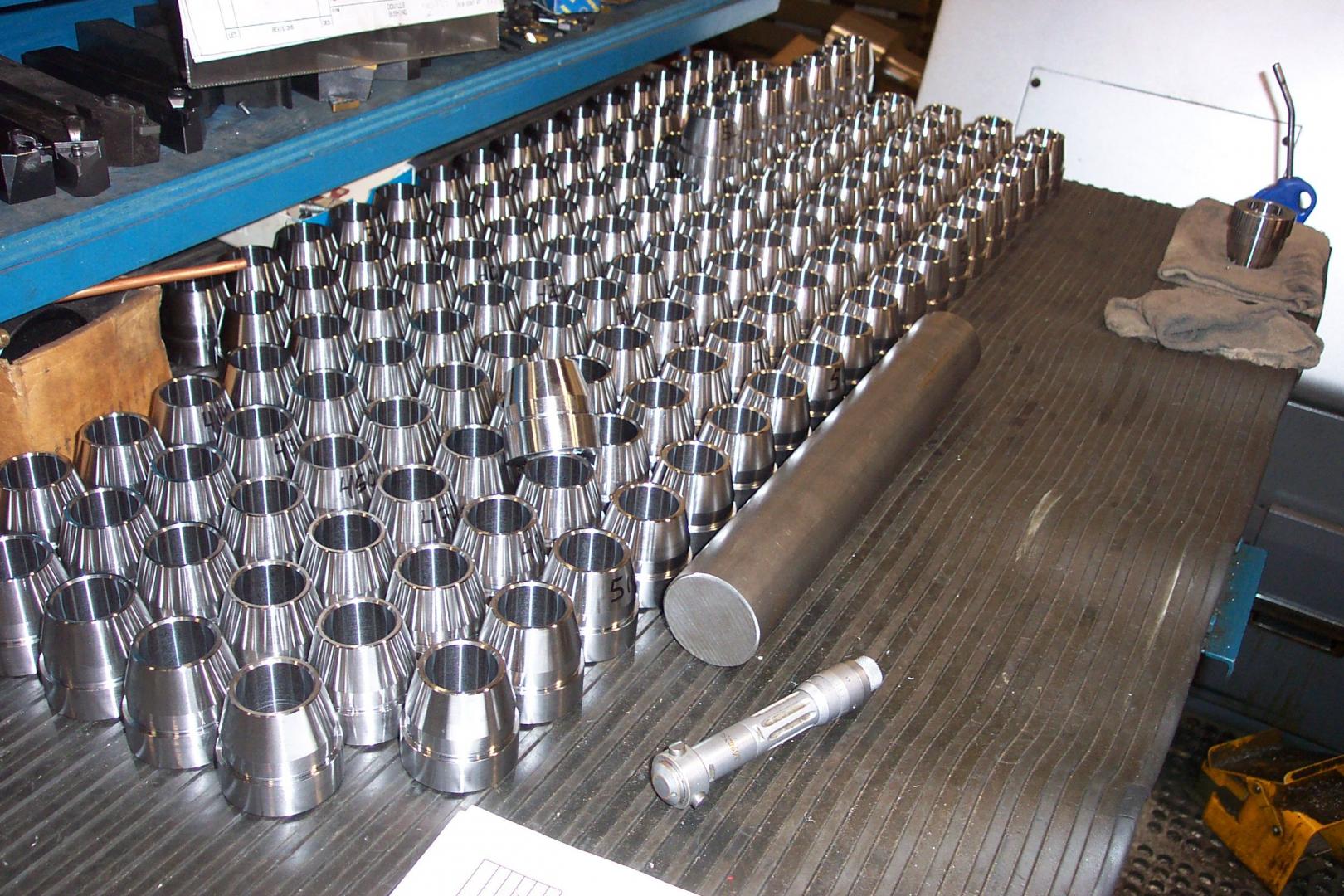 Un établi dans un atelier d'usinage, recouvert de nombreuses pièces cylindriques en métal argenté, d'une tige métallique et d'un outil.