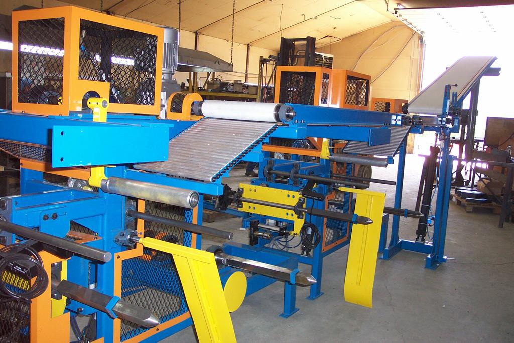 Industrial manufacturing machine with blue and yellow framework, featuring rollers and a conveyor in a warehouse.