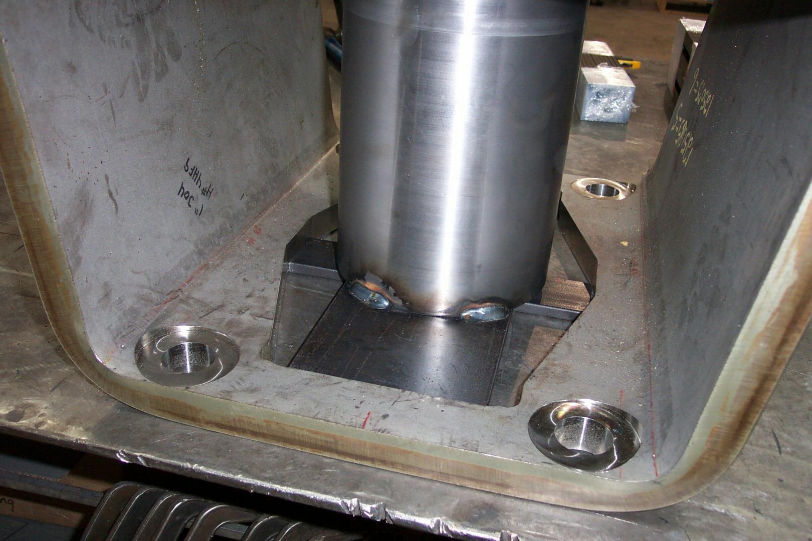 A metal cylinder is welded to a base plate with machined holes inside a larger metal framework in a workshop setting.