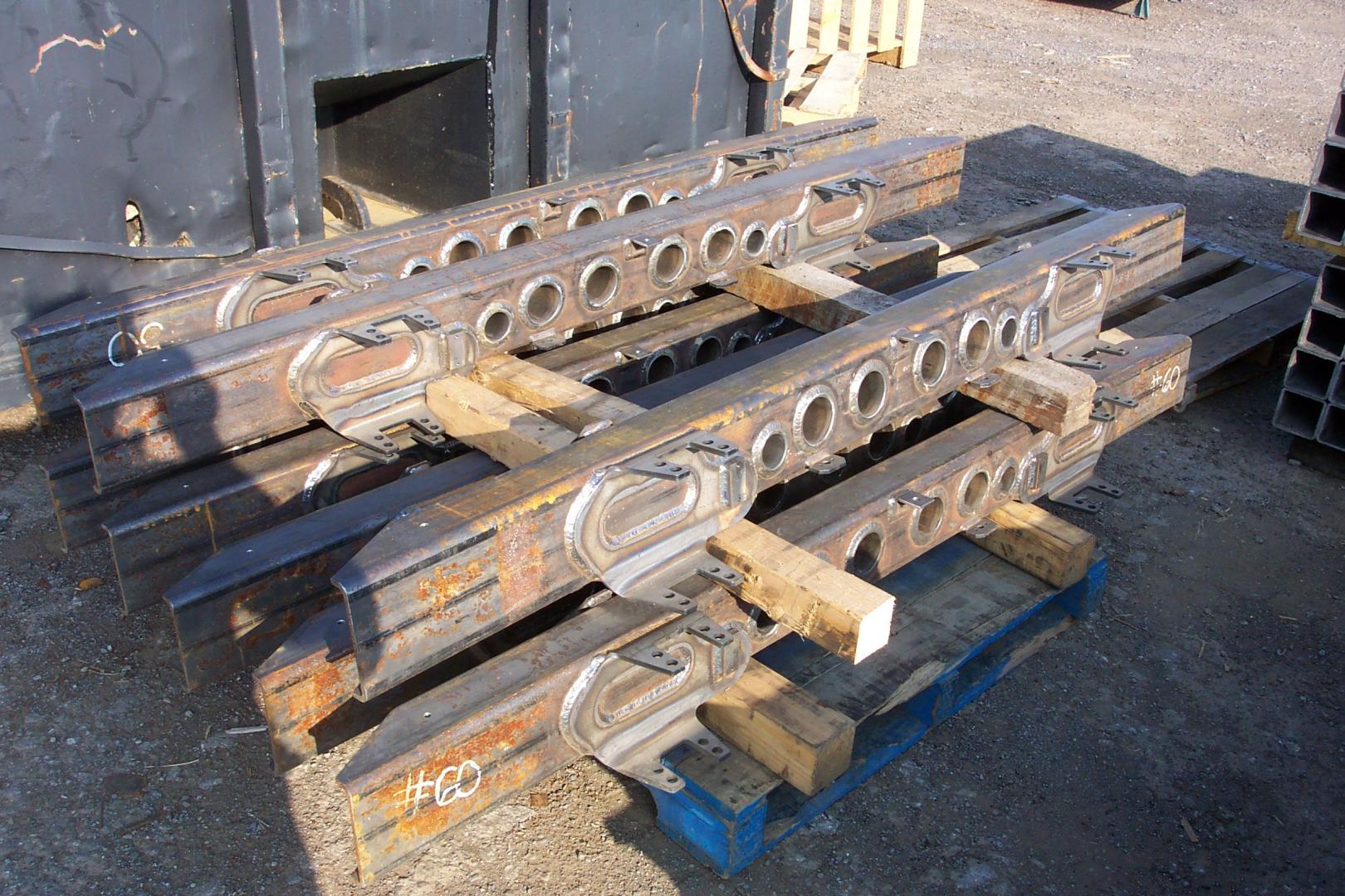 Stacked steel beams with circular cutouts resting on wooden supports on a blue pallet in an industrial yard.
