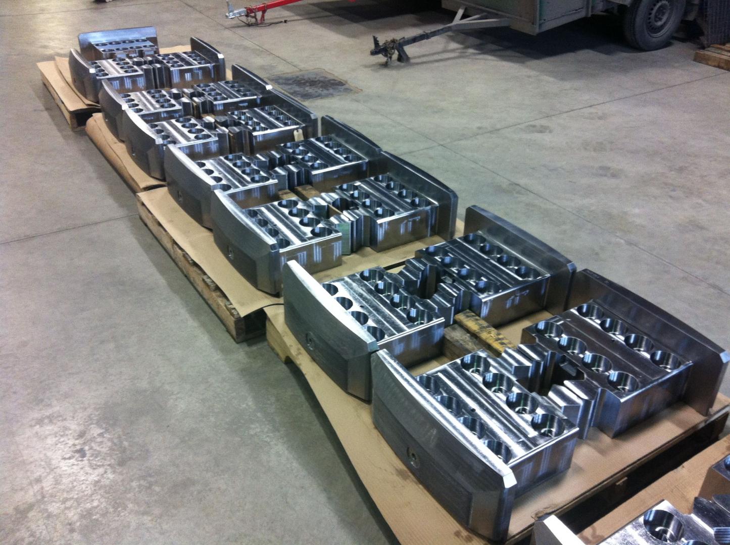 Plusieurs composants métalliques gris, robustes et destinés à un moteur industriel, sont disposés en rangées sur une palette en carton dans un atelier.