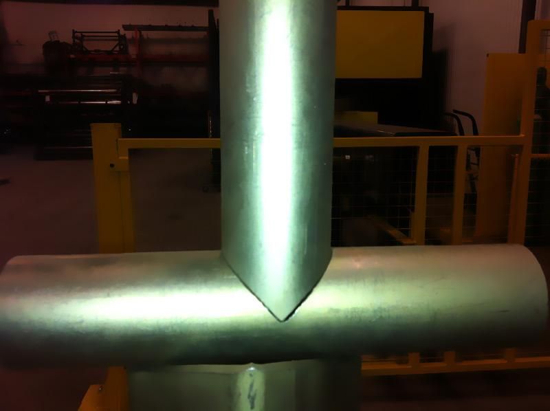 A close-up of a metal pipe T-joint, where a vertical pipe is precision-cut to fit flush against a horizontal pipe.