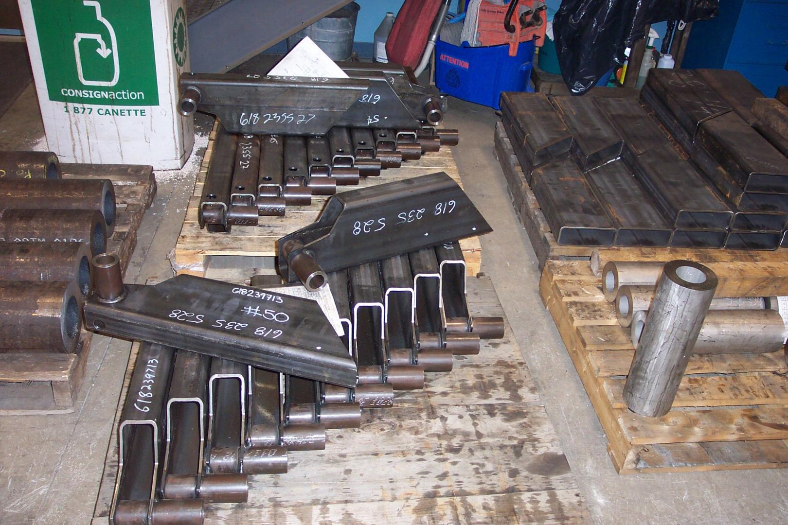 Metal machine parts, including bracket-shaped pieces and hollow cylinders, are stacked on wooden pallets in a warehouse.