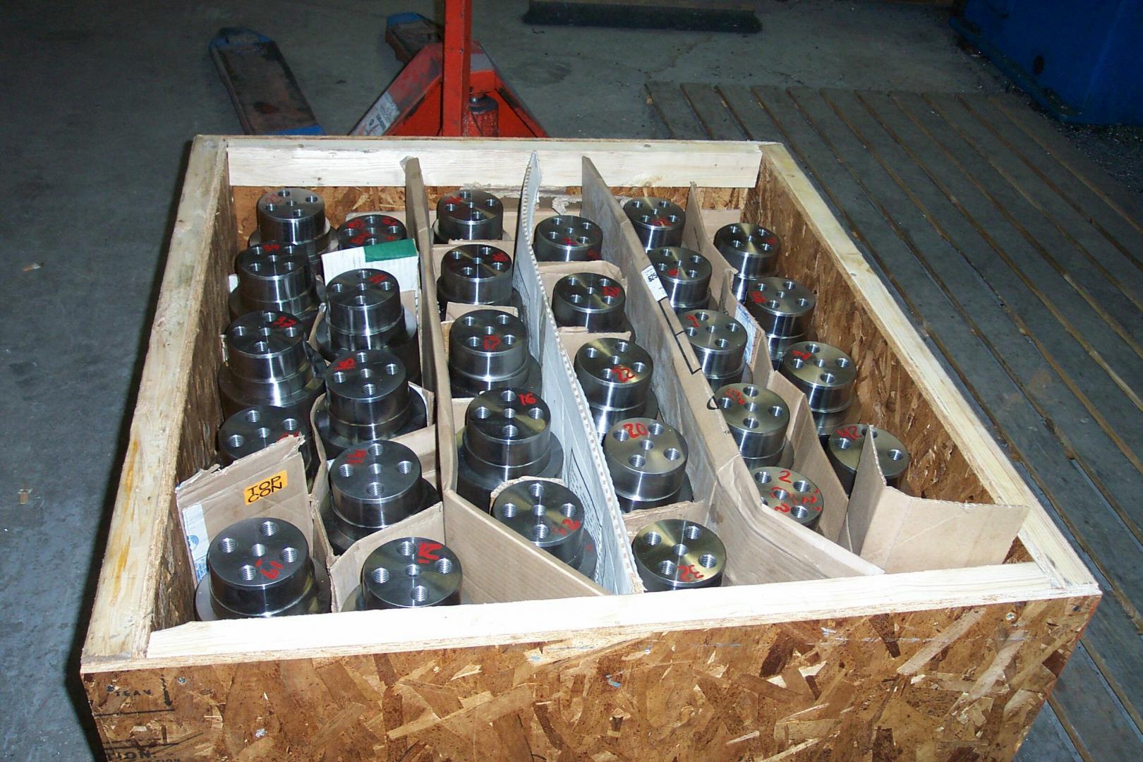 Une caisse en bois remplie de rangées de pièces industrielles cylindriques en métal, séparées par des séparateurs en carton.