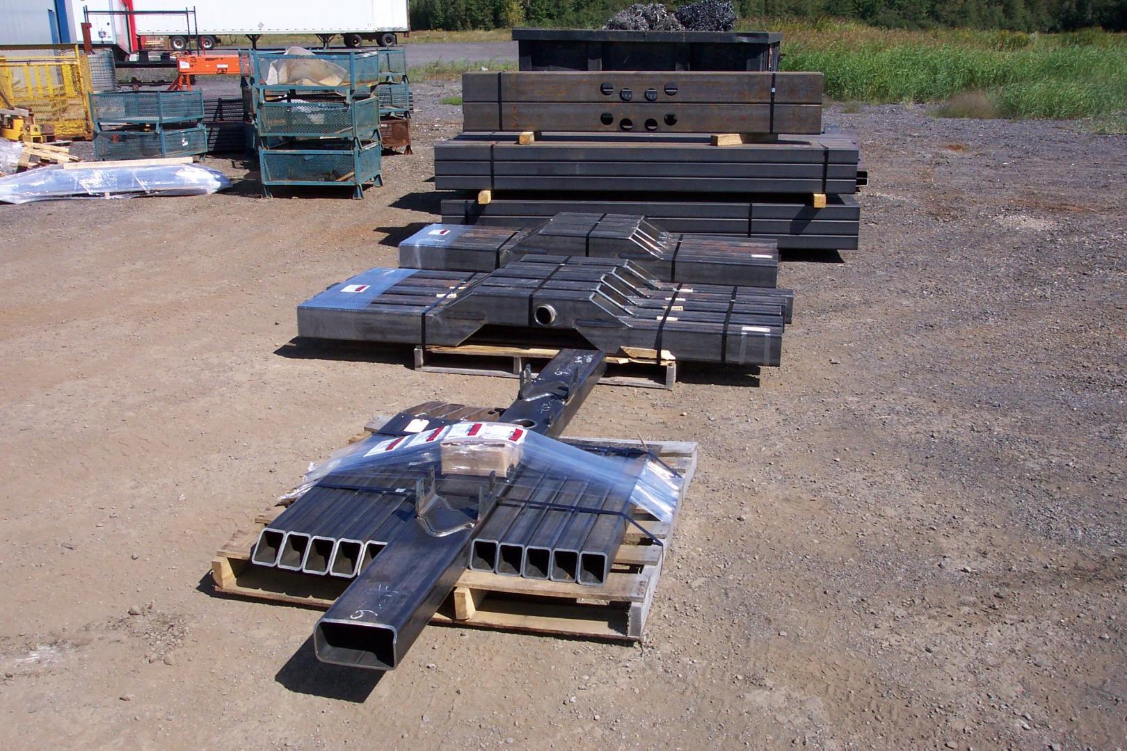 Several stacks of rectangular steel beams and custom fabricated metal components sit on wooden pallets in a gravel yard.