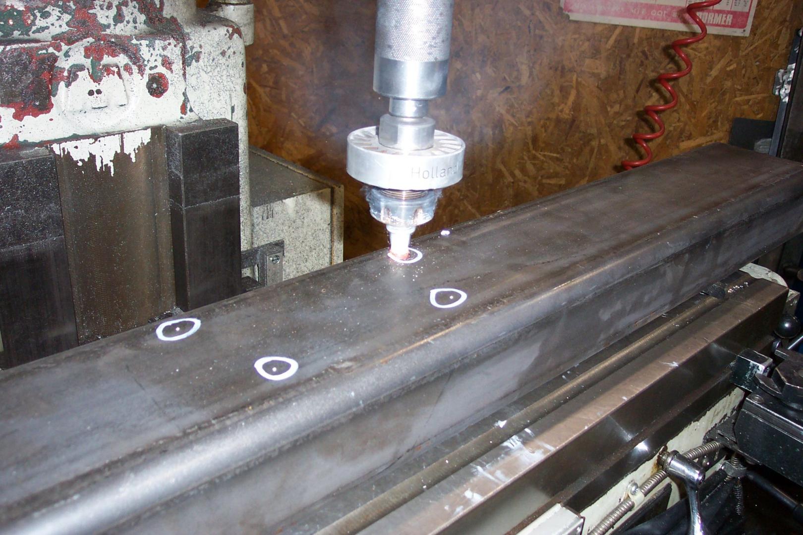 A milling machine tool points toward a metal beam marked with small, white circles indicating drill locations.